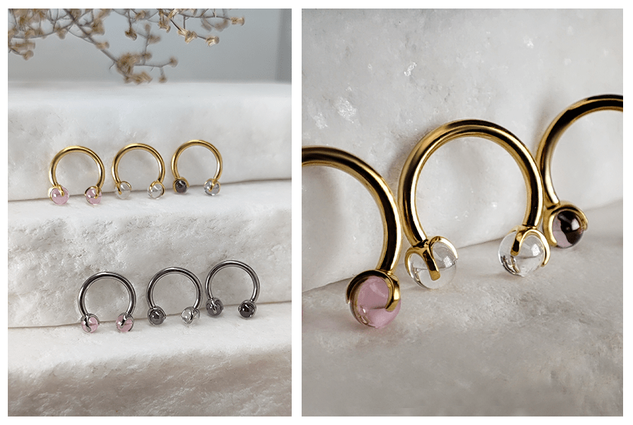 Kolczyki podkówki do piercingu z przezroczystymi kulkami w złotym i stalowym kolorze, ułożone na jasnym kamieniu