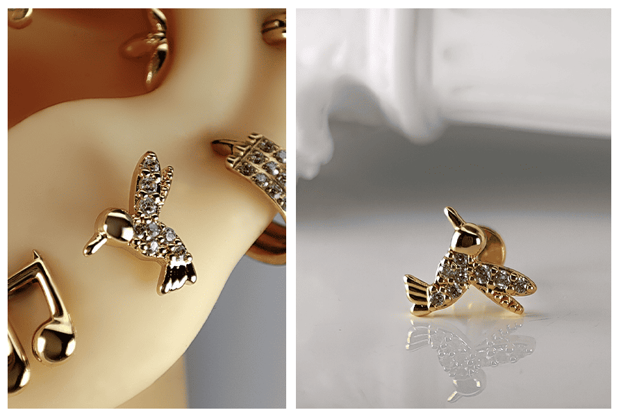 Złoty kolczyk do ucha z motywem kolibra wysadzanym przezroczystymi kryształkami, pokazany w zbliżeniu na silikonowym modelu ucha oraz na jasnym tle z odbiciem