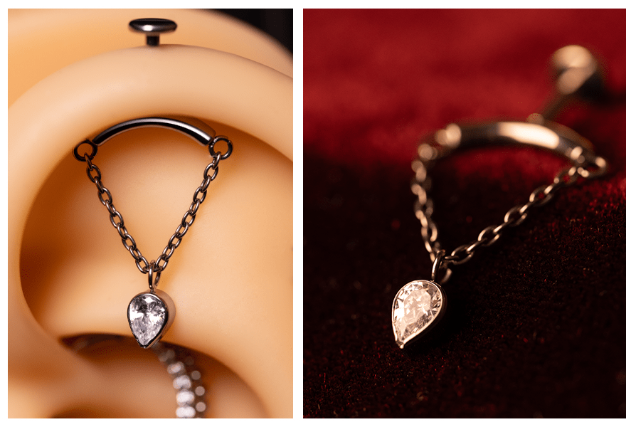 Zbliżenie labretu do vertical helix: łukowaty pręcik, łańcuszek w kształcie V i odwrócona łezka z przezroczystym kamieniem – pokazane na modelu ucha i na czerwonym tle